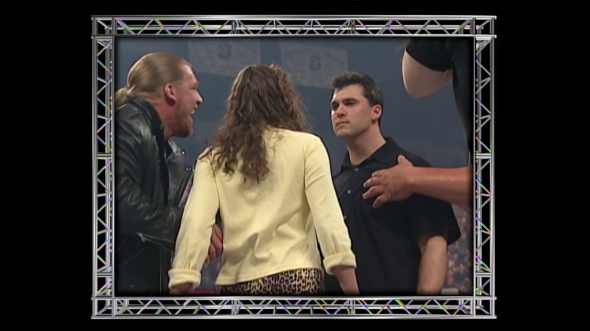 A frame from WWE Raw (March 6, 2000) at 14:12. Three wrestlers are captured in an intense confrontation inside the ring during WWE Raw. A woman in a cream-colored top stands between two men in dark clothing, with metal truss framing visible around the broadcast display.