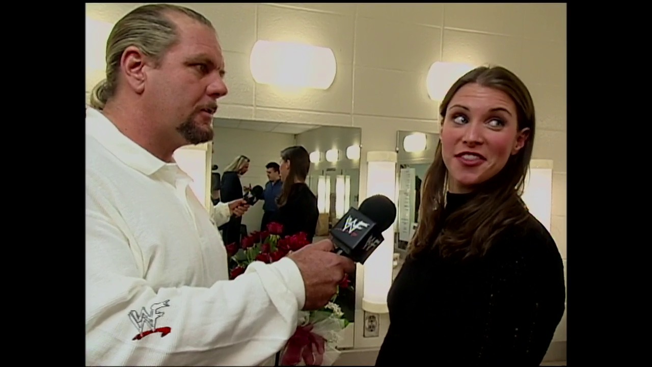 A frame from WWE SmackDown (November 25, 1999) at 37:06. A man in a white WWF jacket holds a microphone while interviewing a woman in a black shirt backstage, with other people visible in the mirror reflection behind them in what appears to be a locker room setting.