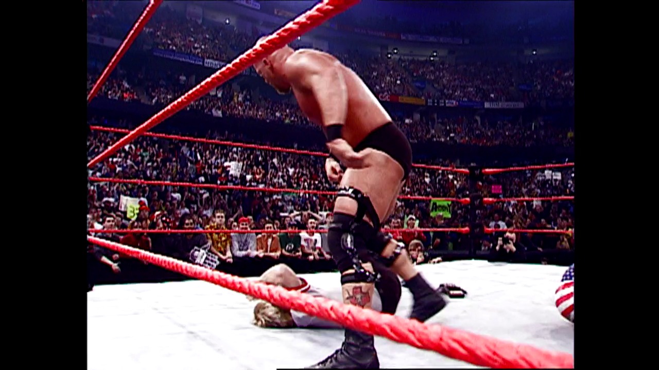 A frame from WWE SmackDown (October 18, 2001) at 1:02. A professional wrestler performs a high-flying move in a WWE ring, leaping over the red ropes with the crowd visible in the background. The arena is filled with spectators, and an American flag is visible at ringside.