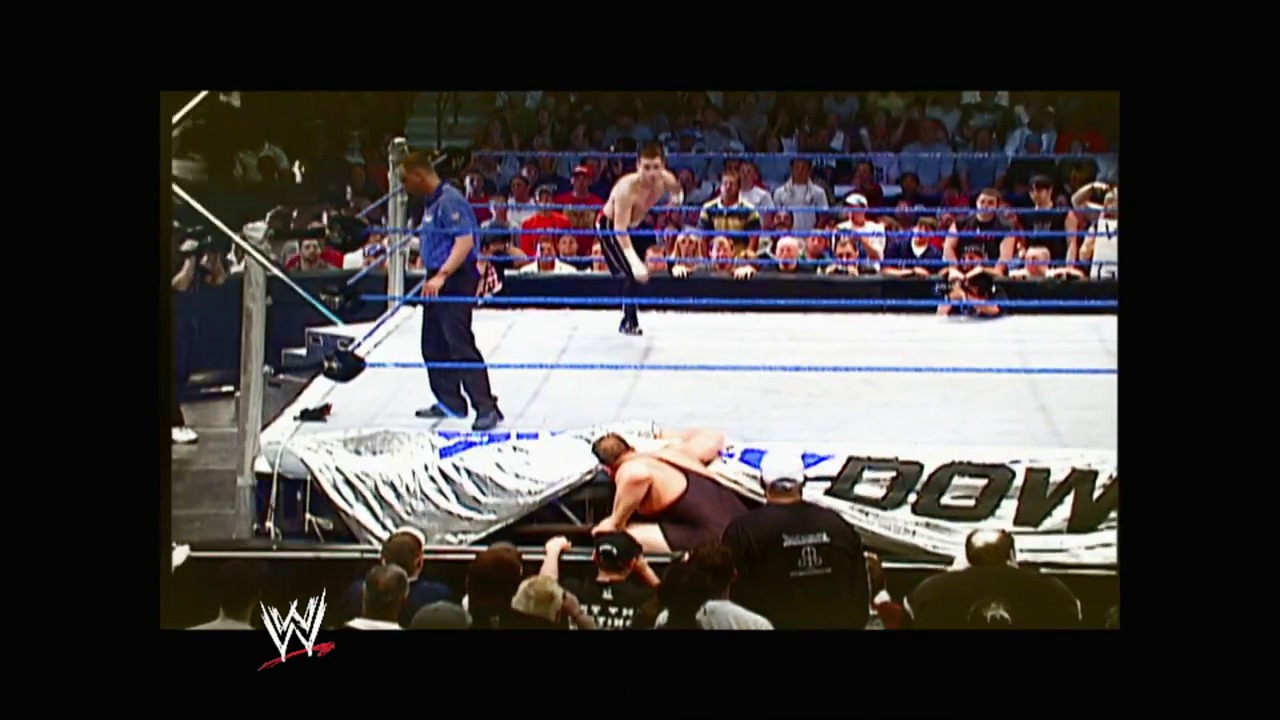A frame from WWE Vengeance (2003) at 1:54:27. A wrestler lies on the mat outside the ring while another stands in the squared circle during a WWE match. The crowd fills the arena seats as the referee and ringside personnel watch the action unfold.