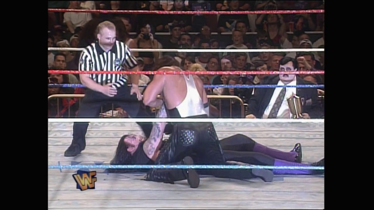 A frame from WrestleMania XII (1996) at 1:11:47. # WrestleMania XII Wrestling Match

A referee in striped shirt stands in the WWF ring as one wrestler delivers an attack to another wrestler on the mat, with a packed crowd visible in the background and the WWF logo displayed at ringside.