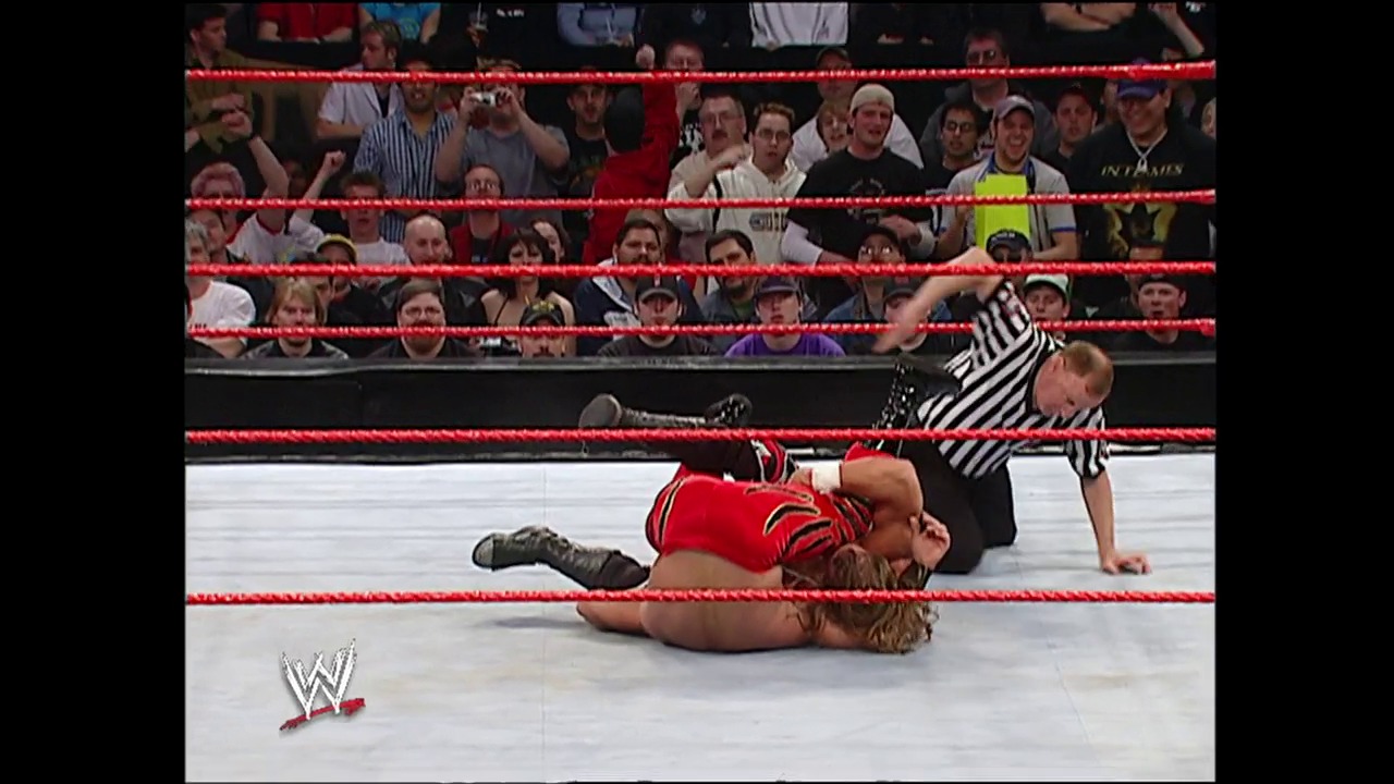 A frame from WWE Backlash (2004) at 2:12:53. A WWE wrestler in red lies on the mat after being knocked down, while a referee in black and white stripes stands over them. The crowd watches from behind the red ropes in the background, with the WWE logo visible in the corner.