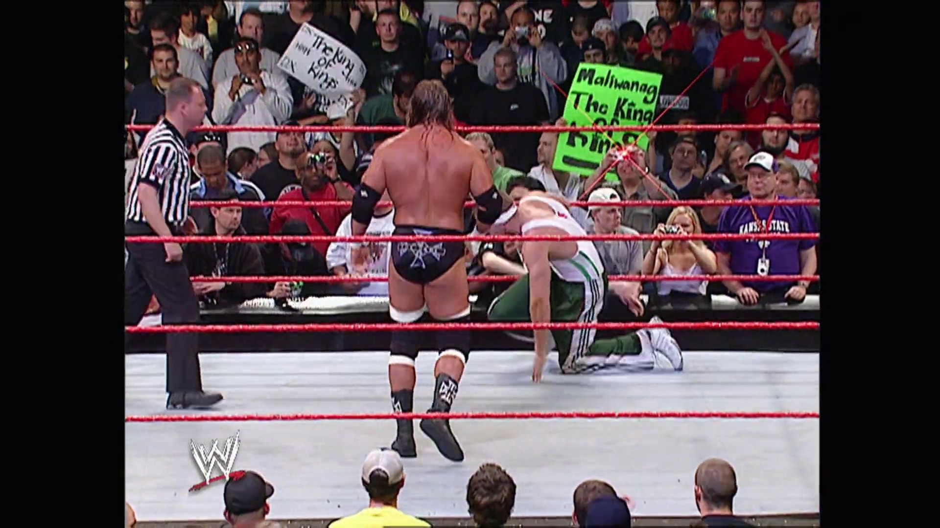 A frame from WWE Raw (May 29, 2006) at 1:12:25. A WWE Raw match in progress shows a shirtless wrestler kneeling in the red-roped ring while an opponent stands nearby, with a referee watching and a packed crowd visible in the background. A green "Malibu" sign is visible among the spectators.
