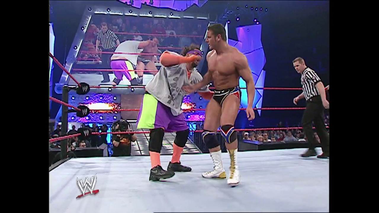 A frame from WWE Raw (November 10, 2003) at 27:40. A WWE wrestler in colorful neon attire engages with a shirtless opponent in the ring while a referee in a striped shirt watches. The purple-lit arena features the WWE logo and crowd visible in the background.