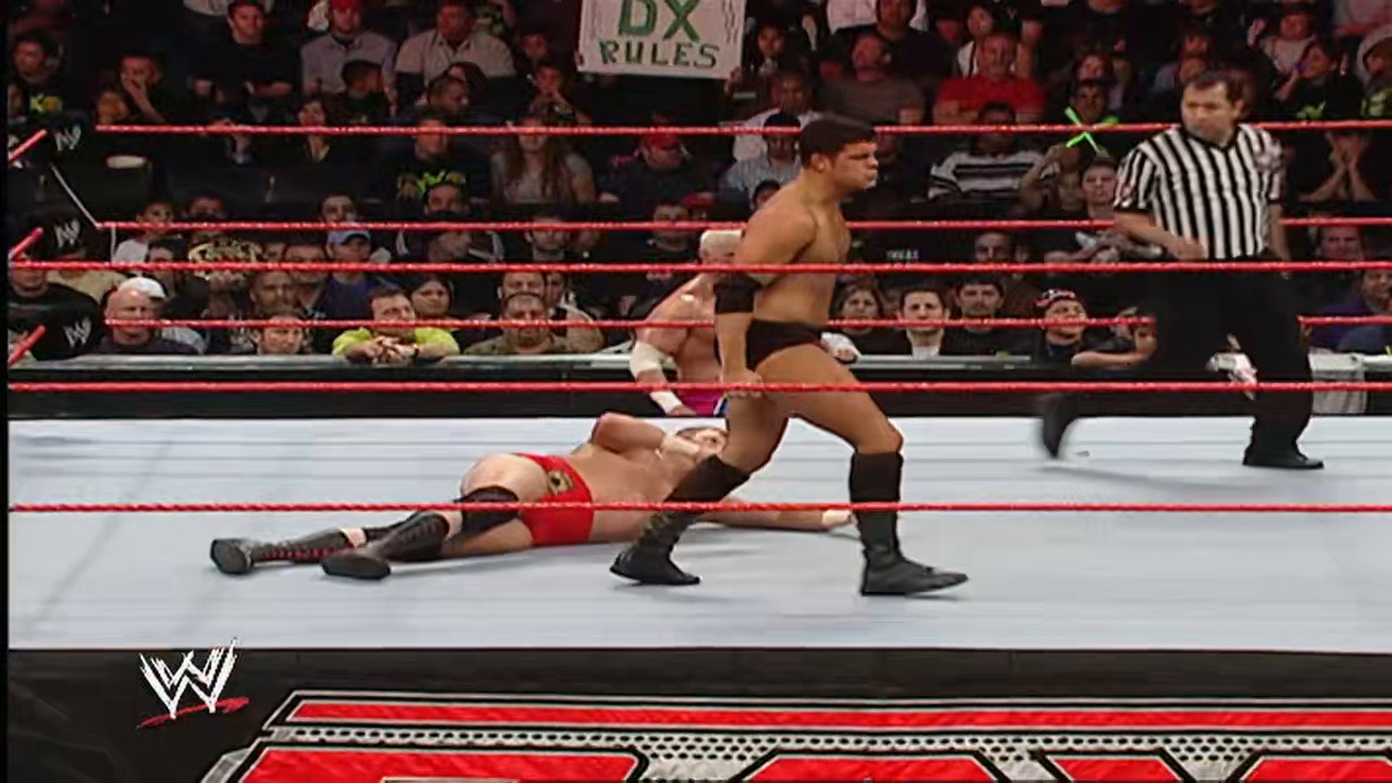 A frame from WWE Raw (November 5, 2007) at 1:07:42. A WWE wrestler in black attire stands over a downed opponent in red in the wrestling ring, with a referee in striped shirt nearby and a packed crowd visible in the background under the red ropes.