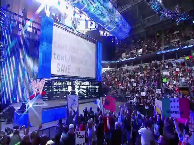 A frame from WWE SmackDown (February 26, 2010) at 1:25. A WWE SmackDown event featuring a packed arena with enthusiastic crowds holding signs, illuminated by blue stage lighting and large video screens displaying match information above the wrestling ring.
