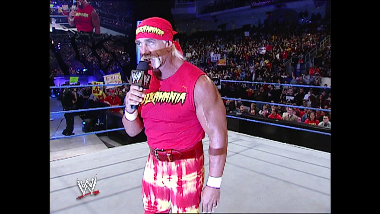 A frame from WWE SmackDown (March 6, 2003) at 36:45. A WWE wrestler in a bright red and yellow bandana and matching sleeveless shirt holds a microphone while standing in the wrestling ring, addressing the crowd during a live broadcast. The arena is filled with spectators in the background, and the WWE logo is visible in the corner of the frame.