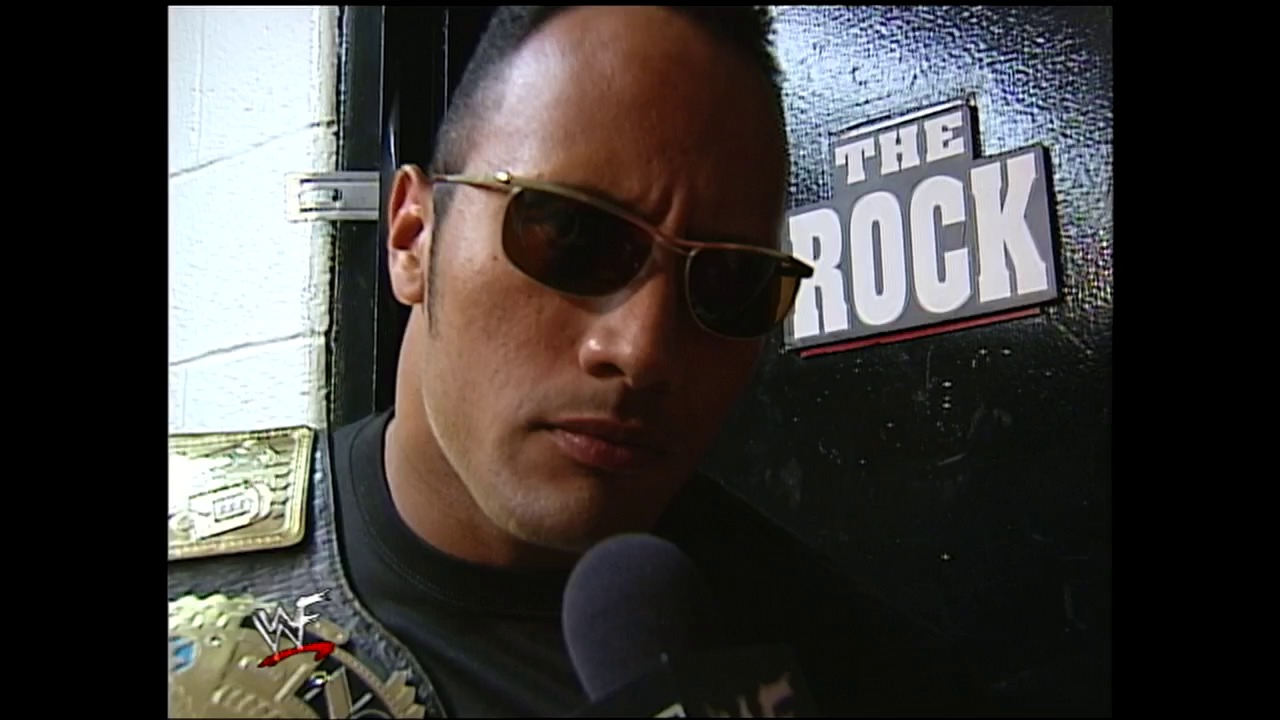 A frame from WWE SmackDown (May 18, 2000) at 1:15:04. A man wearing sunglasses and dark clothing holds a microphone at a WWE event, with "THE ROCK" text visible on a banner behind him. The WWF logo appears in the lower left corner of the frame.