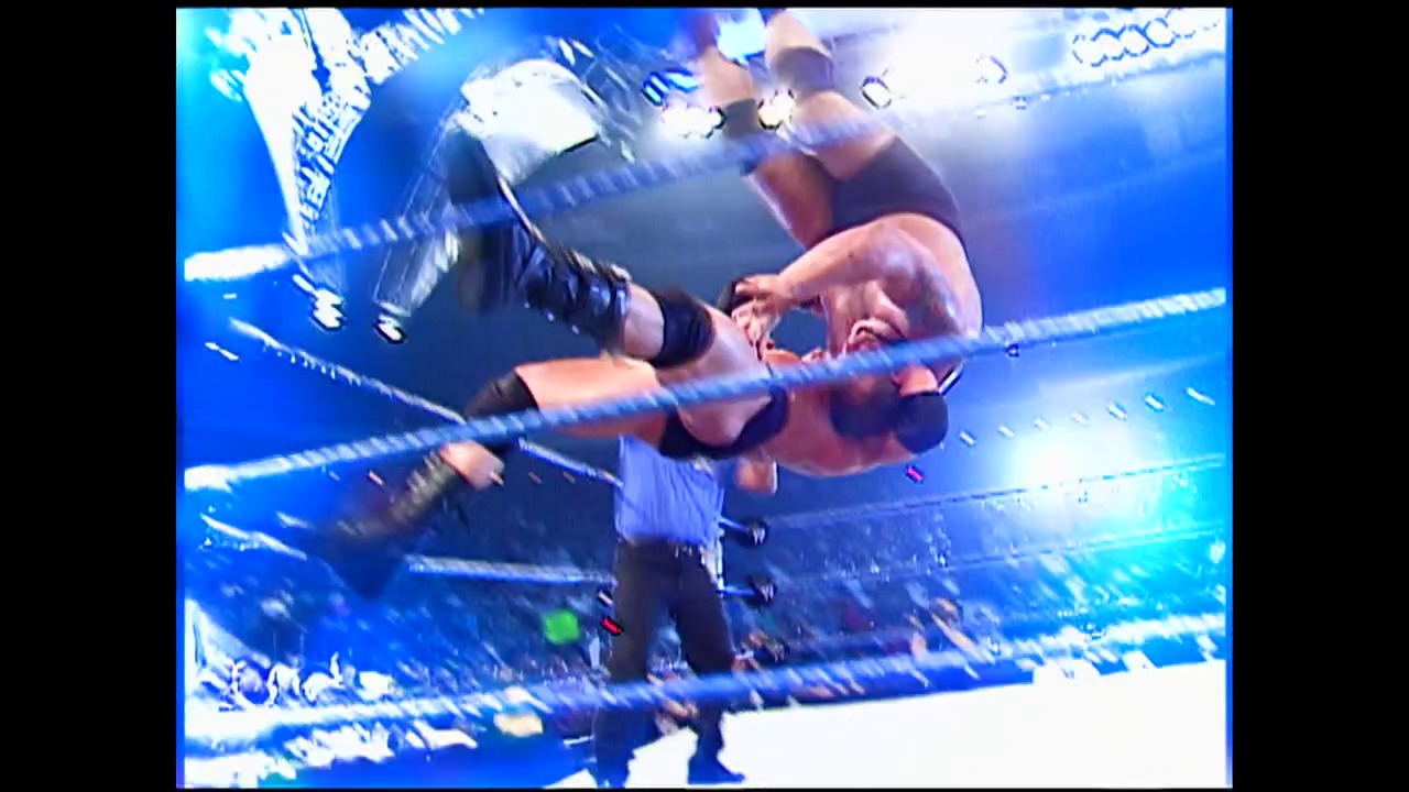 A frame from WWE Vengeance (2003) at 0:45. A wrestler in dark attire performs an aerial maneuver over the blue-roped wrestling ring during an intense WWE match, with dynamic lighting and broadcast graphics visible in the background.