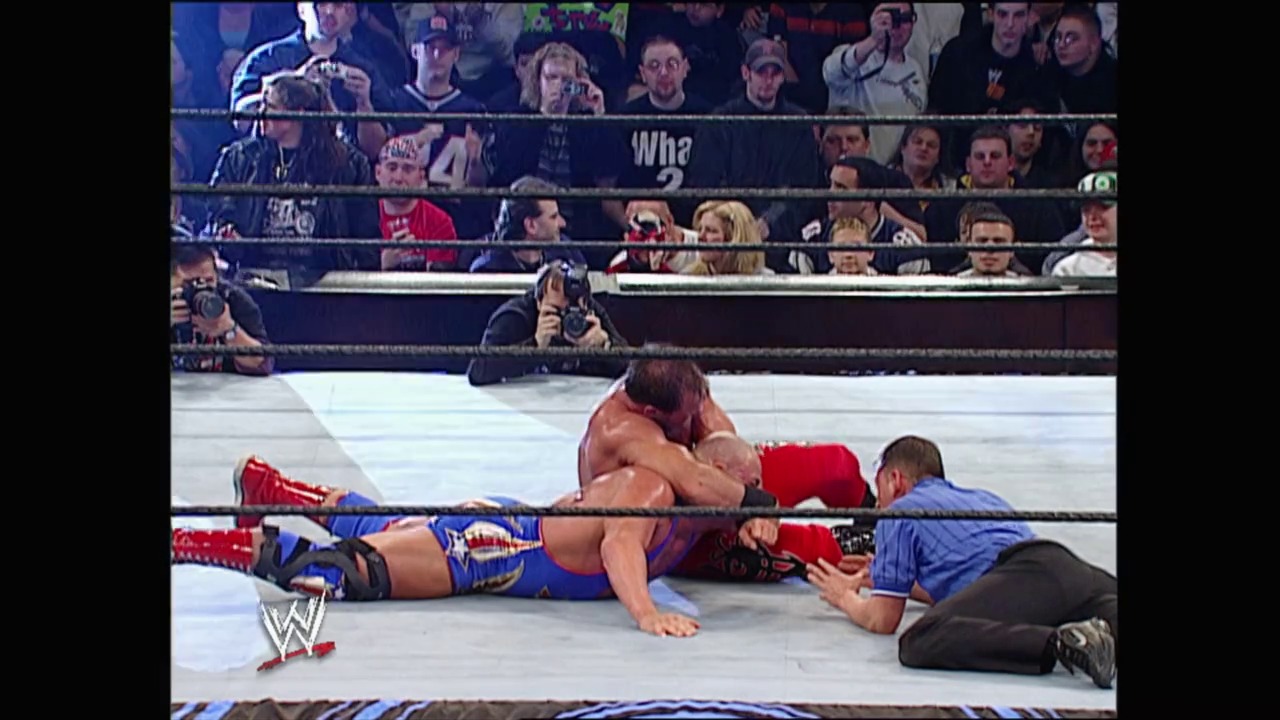 A frame from Royal Rumble (2003) at 1:30:30. A WWE wrestler lies on the canvas during a Royal Rumble match, with a referee checking on him. The crowd is visible in the background behind the ring ropes, and the WWE logo appears in the lower left corner.