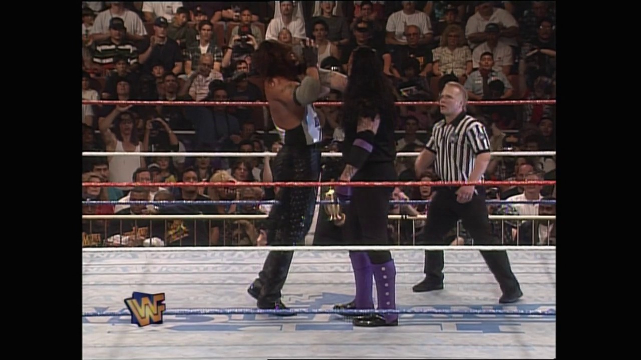 A frame from WrestleMania XII (1996) at 1:12:14. A WWE wrestling match is in progress inside the ring, with two wrestlers facing off while a referee in a striped shirt officiates. The crowd fills the arena seats in the background, and the WWF logo is visible on the ring canvas.