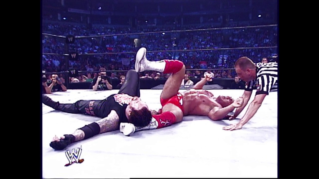 A frame from WWE Raw (July 11, 2002) at 0:31. Two wrestlers are down on the mat during a WWE match, with one in red shorts and the other in black pants. A referee in a striped shirt stands nearby as the crowd and stadium lights fill the background, with the WWE logo visible in the lower left corner.