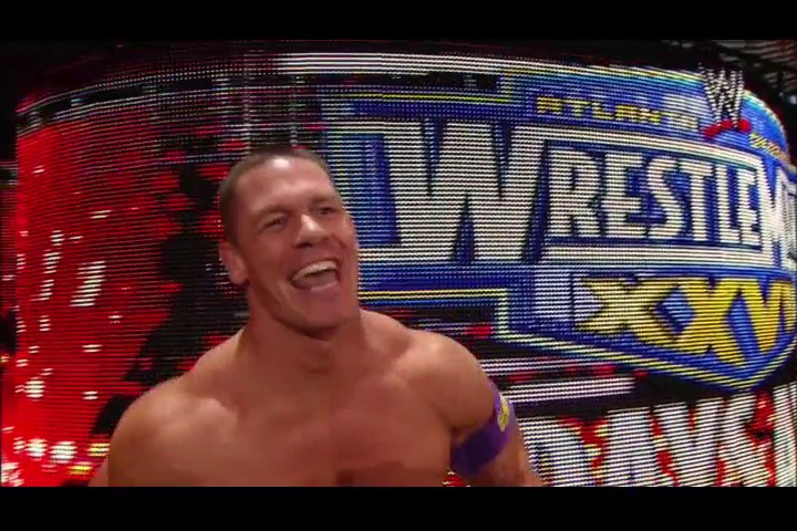 A frame from WWE Raw (March 28, 2011) at 1:40:43. A shirtless male wrestler smiles while standing in front of a large LED display featuring WWE WrestleMania branding in red, blue, and yellow colors at what appears to be a professional wrestling event.