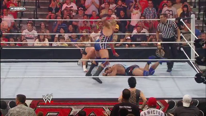 A frame from WWE Raw (October 24, 2011) at 38:19. A WWE wrestler in blue and gold attire delivers an aerial move to an opponent on the mat, while a referee in stripes watches nearby. The packed crowd cheers from the stands as the event branding appears at the bottom of the frame.