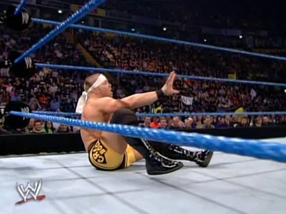 A frame from WWE Rebellion (2002) at 41:45. A WWE wrestler sits on the mat in the ring wearing gold and black attire with a headband, extending his arm toward the camera. The blue-roped ring is surrounded by a packed arena crowd under bright lighting, with the WWE logo visible in the corner.