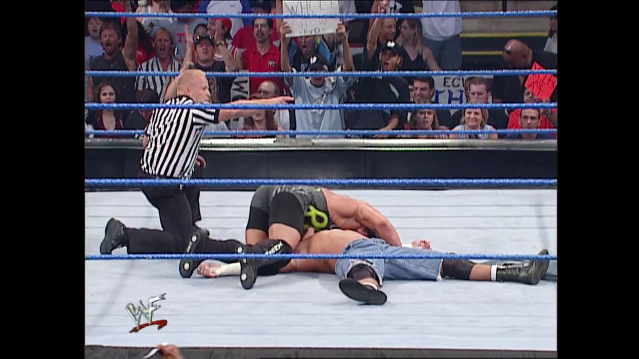 A frame from WWE SmackDown (August 23, 2001) at 12:24. A WWE referee in striped shirt stands over a fallen wrestler on the mat during a SmackDown match, with the crowd visible in the background bleachers and the WWF logo visible on the ring apron.