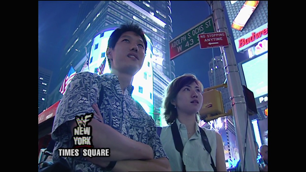 A frame from WWE SmackDown (July 6, 2000) at 1:16:50. Two people stand in Times Square at night, looking upward at the illuminated skyscrapers and digital displays surrounding them. The WWE logo and "NEW YORK" text appear in the lower left corner, identifying this as a WWE SmackDown broadcast location shot.