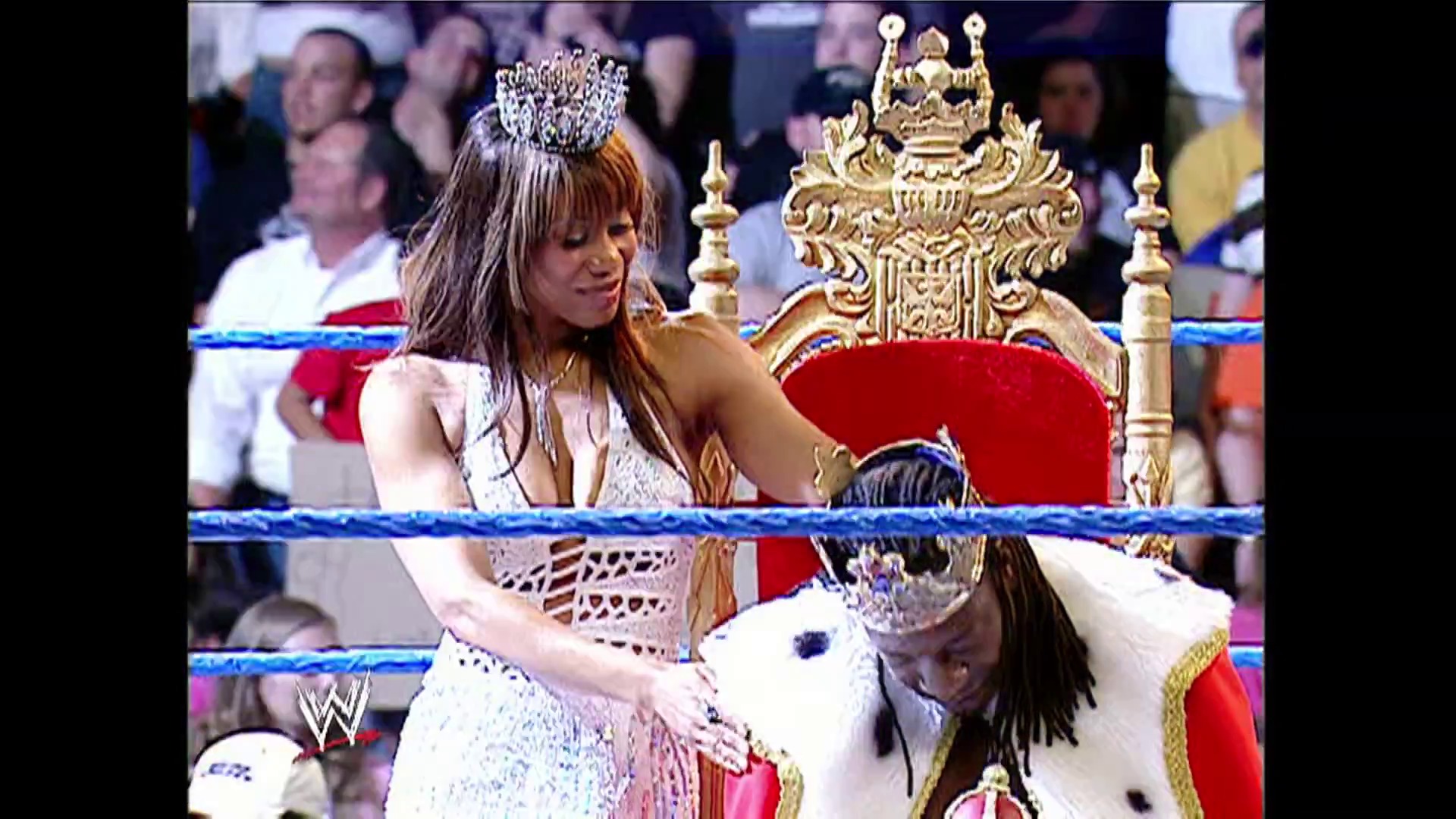 A frame from WWE SmackDown (June 2, 2006) at 1:10:41. A woman in an ornate crown and white outfit sits on an elaborate gold throne with red velvet in a WWE wrestling ring, surrounded by cheering spectators and blue ring ropes.
