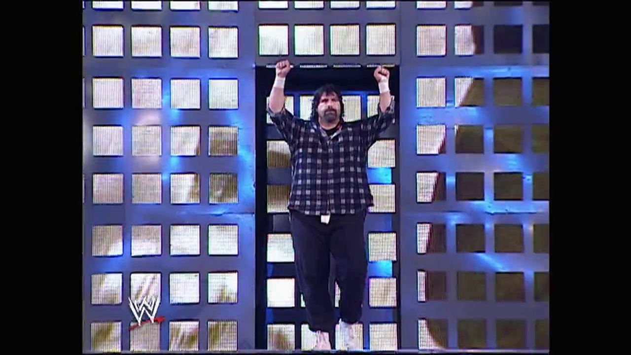 A frame from WrestleMania 22 (2006) at 1:06:24. A wrestler stands triumphantly with both fists raised on a WWE entrance stage, wearing a checkered shirt and dark pants. Behind him is an elaborate backdrop featuring illuminated window-like panels arranged in a grid pattern with blue lighting effects.