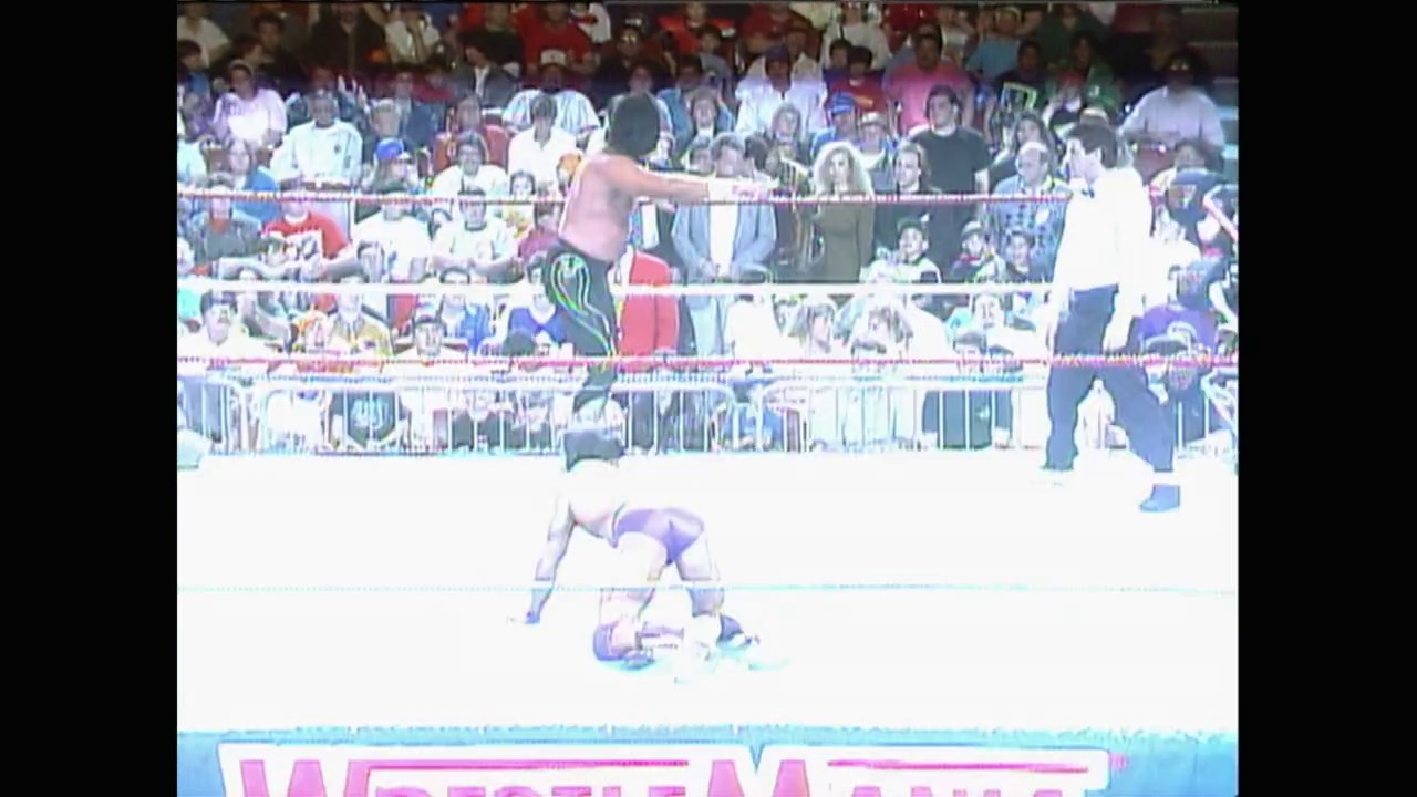 A frame from WrestleMania VII (1991) at 1:00:10. A wrestler in purple lies on the mat while another in red stands above in the ring, with a packed crowd visible in the stands behind the ropes and WrestleMania branding visible at the bottom of the frame.