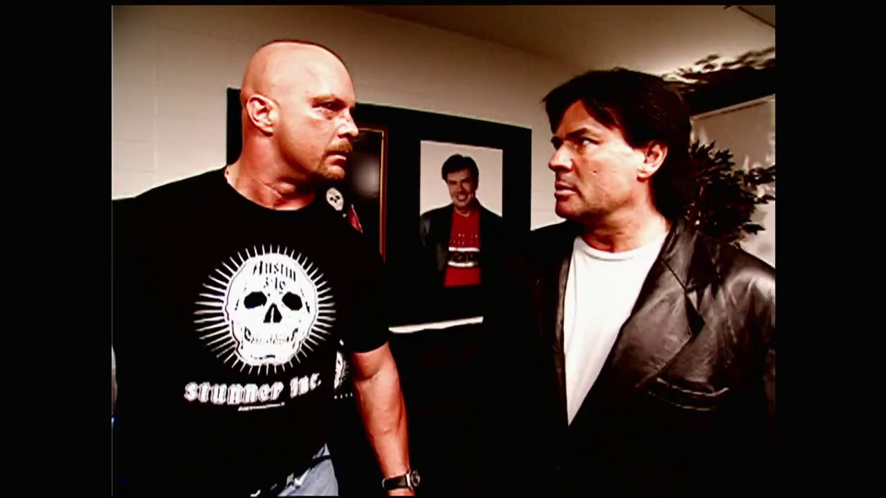 A frame from WWE Bad Blood (2003) at 0:34. Two men face each other in an indoor setting, with the man on the left wearing a black skull-graphic t-shirt and the man on the right in a brown leather jacket over a white shirt. A third person is visible in the background doorway wearing red.