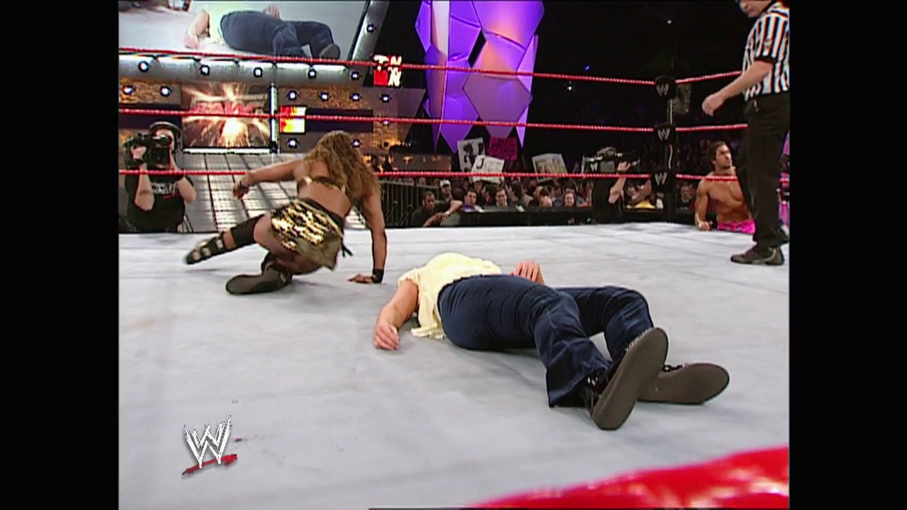 A frame from WWE Raw (February 17, 2003) at 26:58. A WWE wrestler wearing a championship belt performs a move over a fallen opponent in the ring, with a referee nearby and the crowd visible in the background. The red ropes and WWE branding are visible around the wrestling ring.