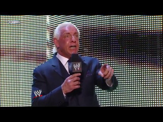A frame from WWE Raw (January 28, 2008) at 1:05:44. A bald man in a dark blue suit holds a WWE microphone while speaking on stage, with an illuminated LED screen backdrop behind him displaying the WWE logo.