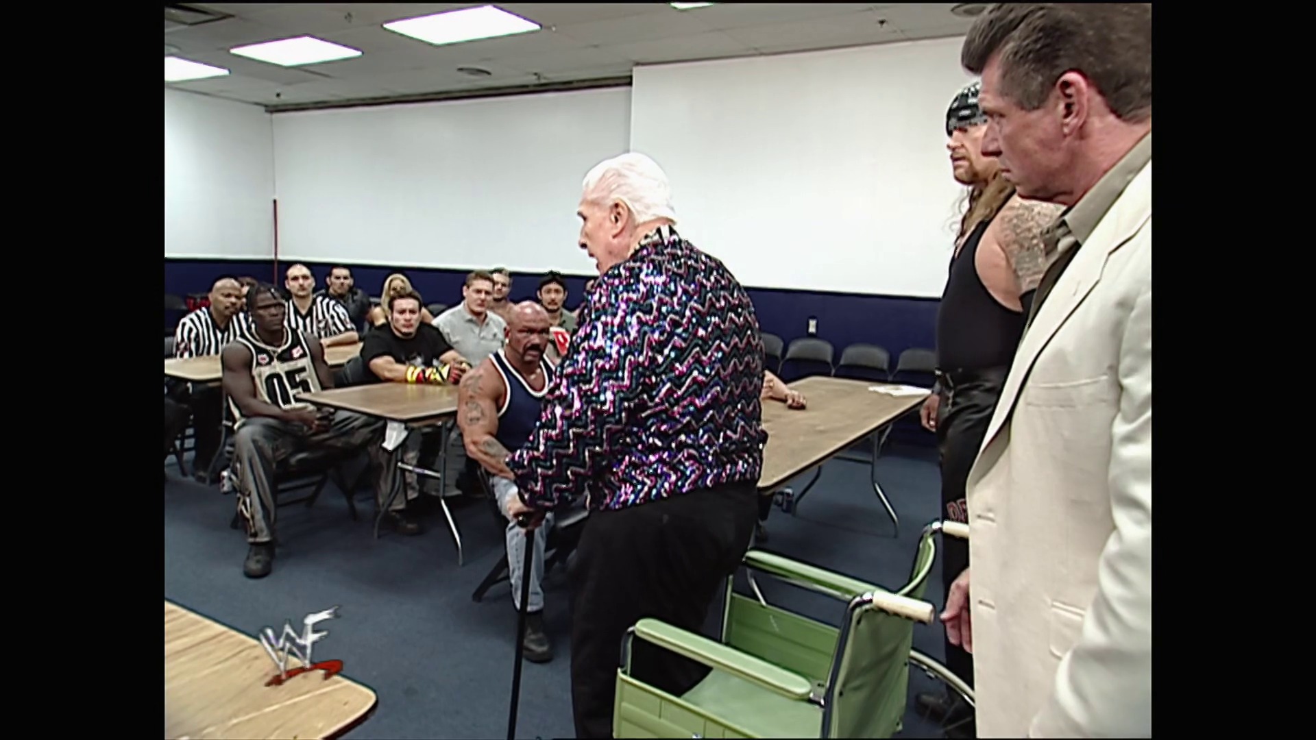 A frame from WWE Raw (July 16, 2001) at 1:15:26. # WWE Raw Visual Description

A WWE segment takes place in a small gymnasium with an audience seated around tables. An older man in a colorful patterned shirt stands center, facing several wrestlers and officials in a casual meeting setup rather than an in-ring confrontation.