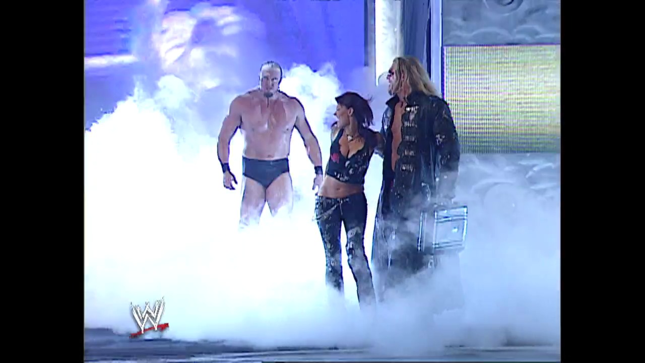 A frame from WWE Raw (June 27, 2005) at 13:56. Three wrestlers emerge through dramatic white smoke and pyrotechnics on the WWE Raw stage, with the shirtless male performer on the left and two other wrestlers in wrestling attire beside him, while the WWE logo appears in the lower left corner.