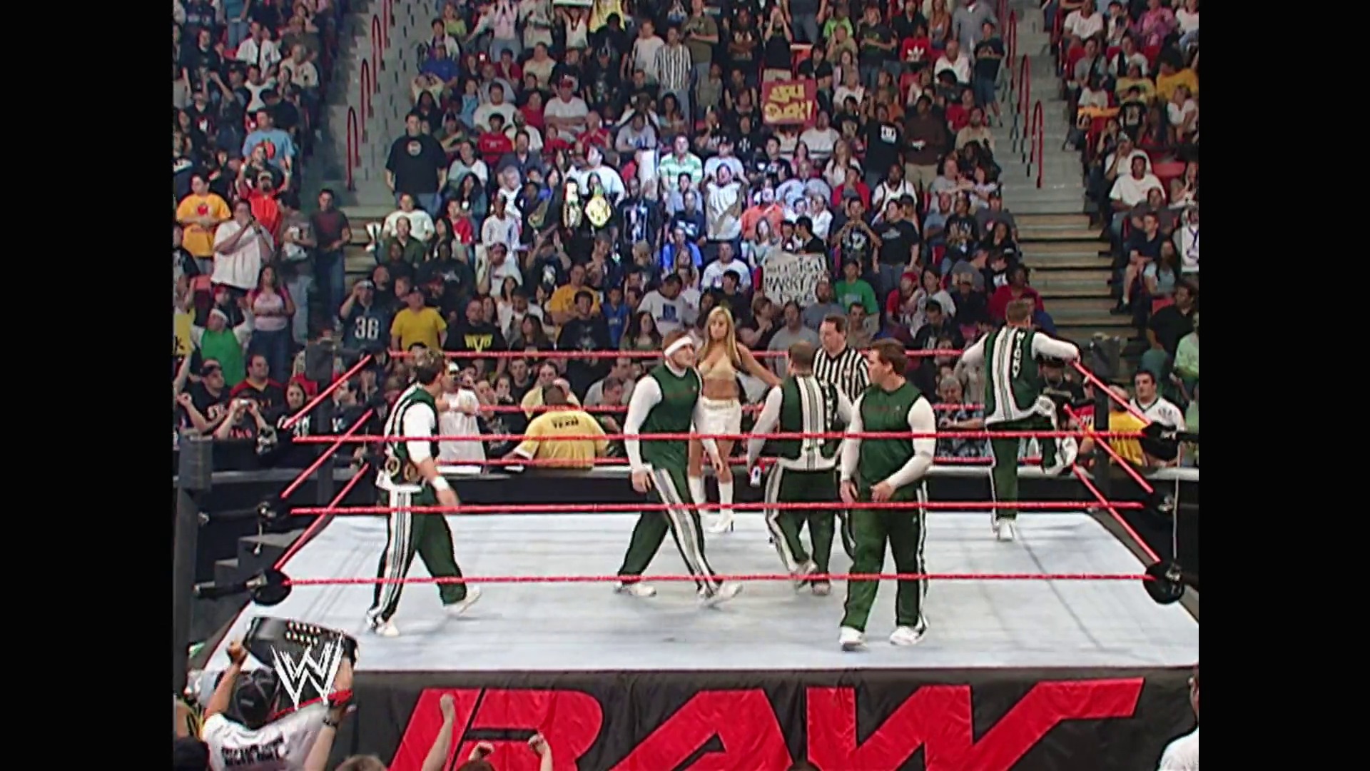 A frame from WWE Raw (May 22, 2006) at 1:14:28. A WWE Raw match features wrestlers in green and white attire in the red-roped ring, with a referee present and a large crowd visible in the background. The WWE logo is prominently displayed at the bottom of the frame.