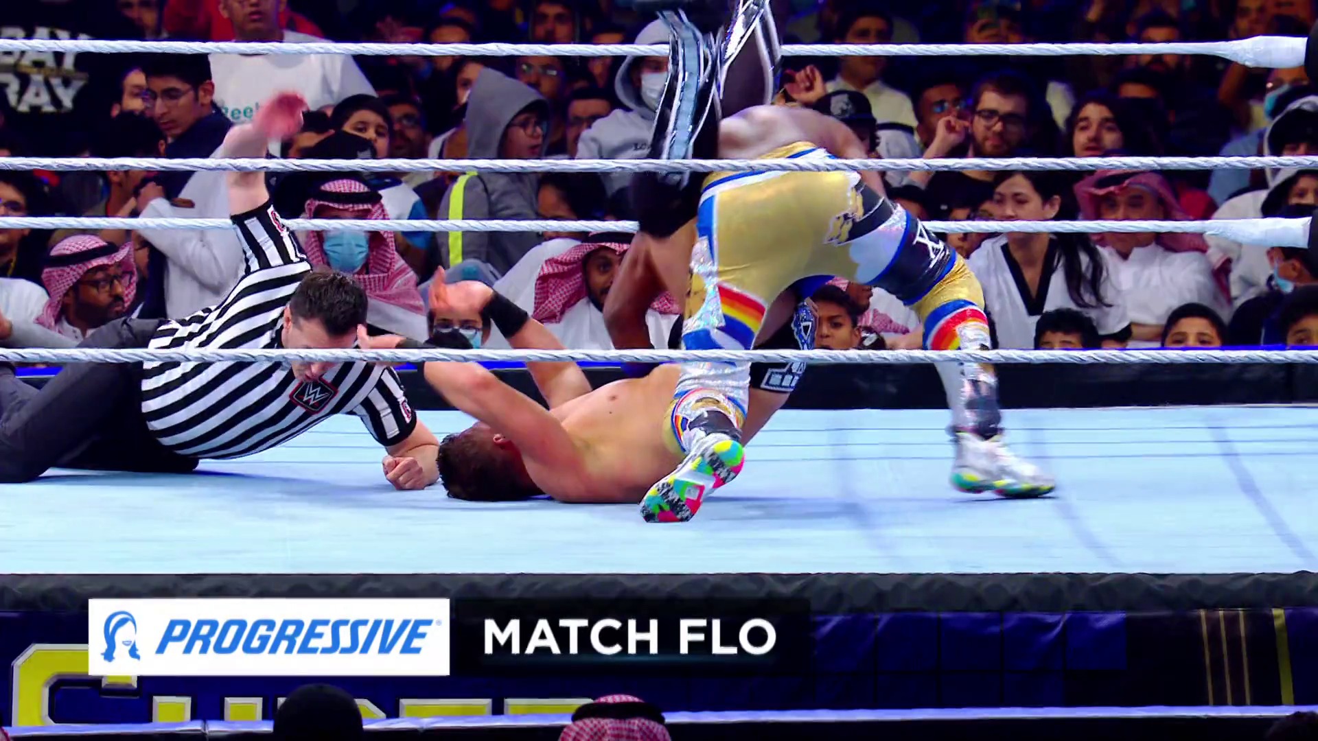 A frame from WWE SmackDown (February 28, 2020) at 1:01:15. A WWE wrestler in colorful yellow and blue attire lies on the mat during a match, while a referee in black-and-white striped shirt observes. The crowd fills the background behind the ring ropes, with "Progressive" and "Match Flo" graphics visible at the bottom.