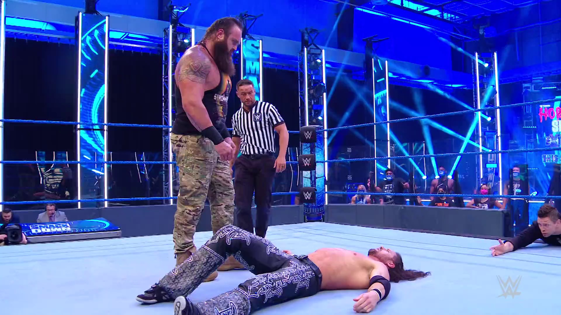 A frame from WWE SmackDown (July 17, 2020) at 47:41. # WWE SmackDown Wrestling Match

A muscular wrestler in camouflage pants stands over a fallen opponent on the mat, while a referee in black and white stripes monitors the action. The ring is surrounded by blue neon lighting and digital screens, with audience members visible at ringside.