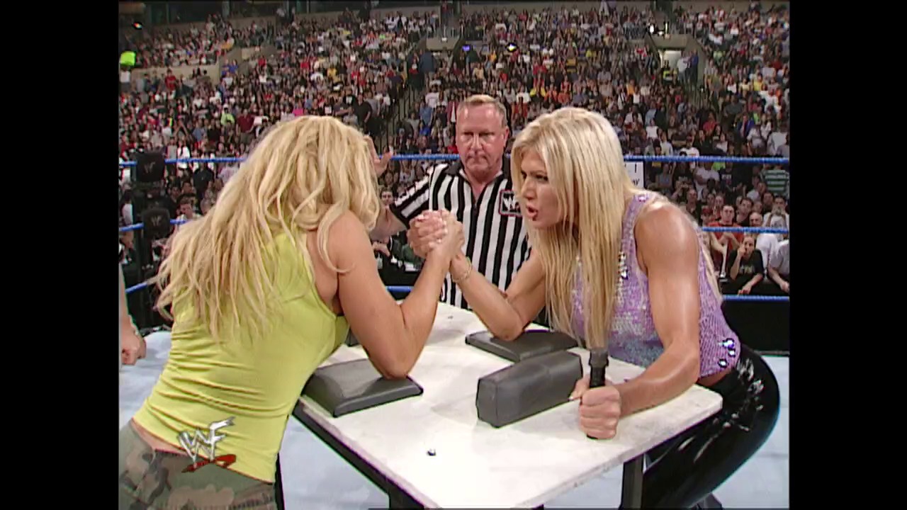 A frame from WWE SmackDown (July 19, 2001) at 59:58. Two blonde female wrestlers compete in an arm-wrestling match at the ring, with a referee in striped shirt overseeing the competition. A packed stadium crowd fills the background of the WWE SmackDown event.