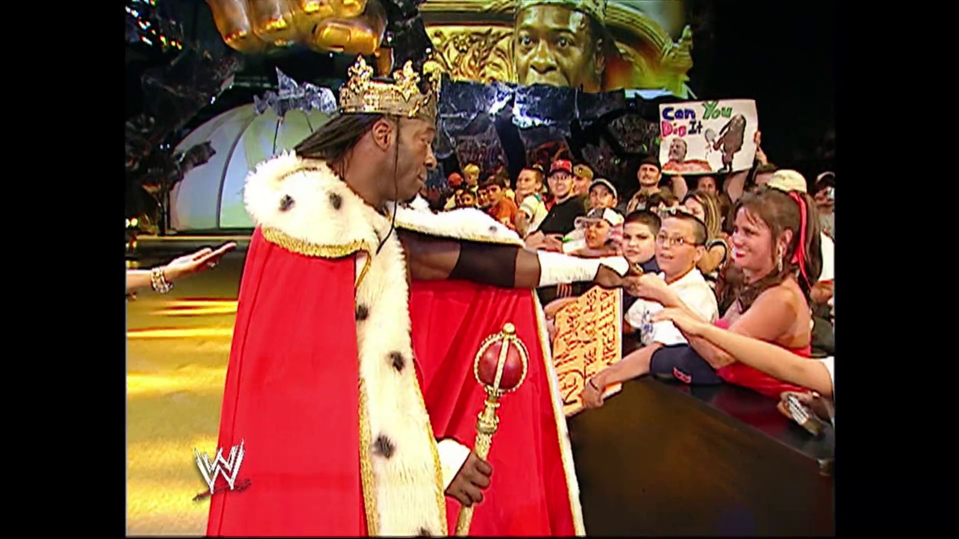 A frame from WWE SmackDown (July 21, 2006) at 1:14:12. A WWE performer dressed in royal attire with a red cape and crown interacts with children at ringside during a SmackDown event, while the crowd watches from the background in an ornate arena setting.