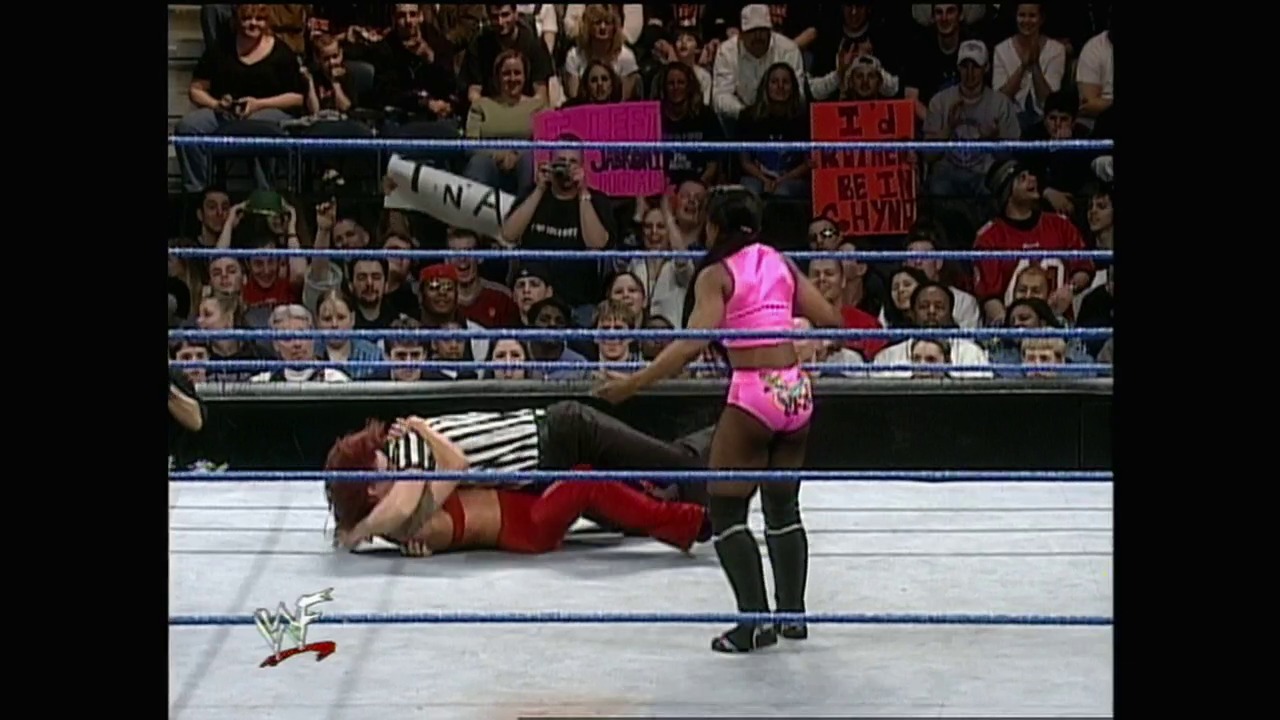 A frame from WWE SmackDown (March 23, 2000) at 1:06:31. # WWE SmackDown Wrestling Match

A wrestler in pink attire stands in the ring while an opponent in red lies on the mat. The crowd fills the stands behind the blue-roped wrestling ring, with visible fan signs and the WWF logo displayed at ringside.