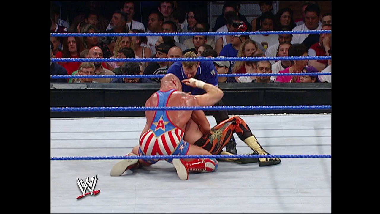 A frame from WWE SmackDown (September 2, 2004) at 1:14:24. A WWE wrestler in red and white patriotic attire applies a submission move to an opponent on the blue-roped ring, while the crowd watches from the stands. The WWE logo appears in the bottom left corner of the broadcast.