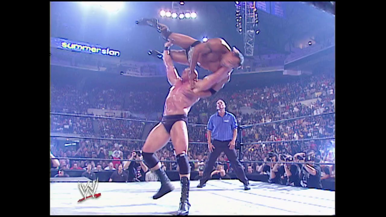 A frame from Summerslam (2002) at 2:40:56. A shirtless wrestler lifts his opponent overhead in the wrestling ring at SummerSlam 2002, with a referee in a striped shirt observing nearby. The packed arena is visible in the background beneath bright stadium lights, with the SummerSlam logo displayed on the arena signage.