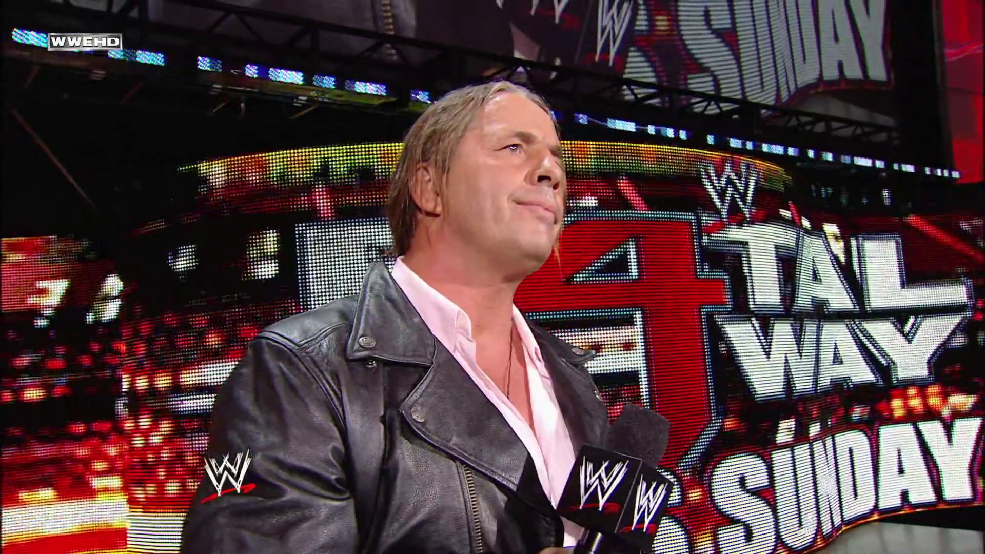 A frame from WWE Raw (June 14, 2010) at 13:20. A WWE performer stands in the ring wearing a black leather jacket over a pink shirt, gazing upward with a composed expression. Behind him, a large LED screen displays the WWE Raw branding and colorful graphics typical of the wrestling event production.