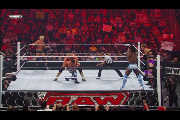 A frame from WWE Raw (September 26, 2011) at 1:04:36. A WWE Raw match in progress features wrestlers in the ring with a referee, surrounded by an energetic crowd illuminated in red lighting. The WWE Raw branding is visible on the ring apron, and the venue is filled with fans holding signs.