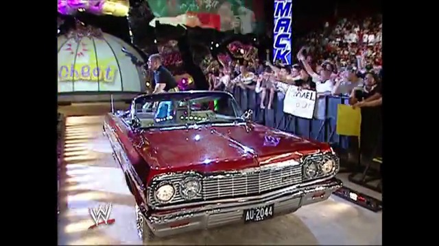 A frame from WWE SmackDown (April 1, 2005) at 0:44. A vintage red convertible car is displayed in a WWE wrestling arena surrounded by enthusiastic crowds. The vehicle features classic 1960s styling with distinctive chrome detailing and is positioned on the illuminated floor in front of packed bleachers.
