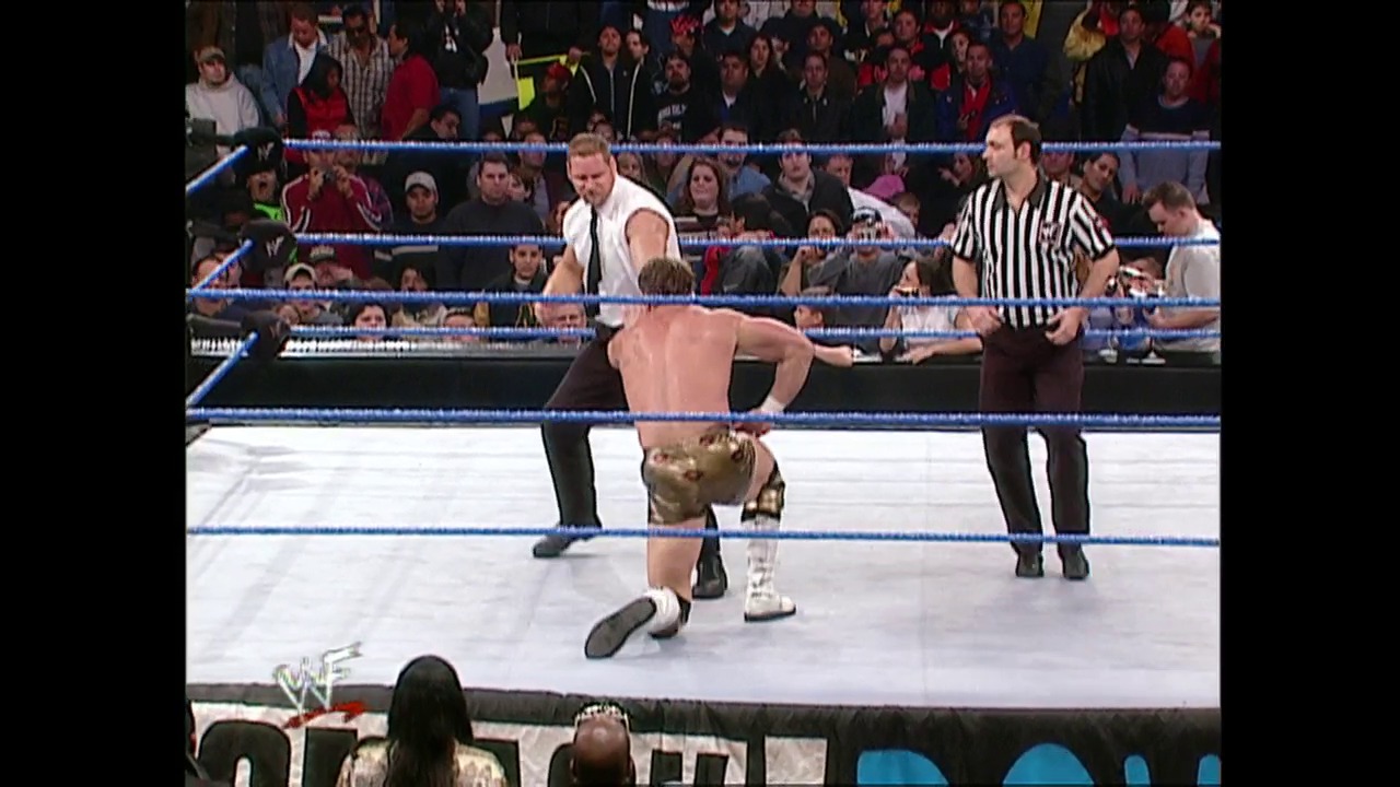 A frame from WWE SmackDown (January 4, 2001) at 1:04:33. A WWE wrestler performs a move in the ring while another competitor and a referee in striped shirt watch. The crowd fills the arena stands behind the blue-roped ring during this SmackDown event.