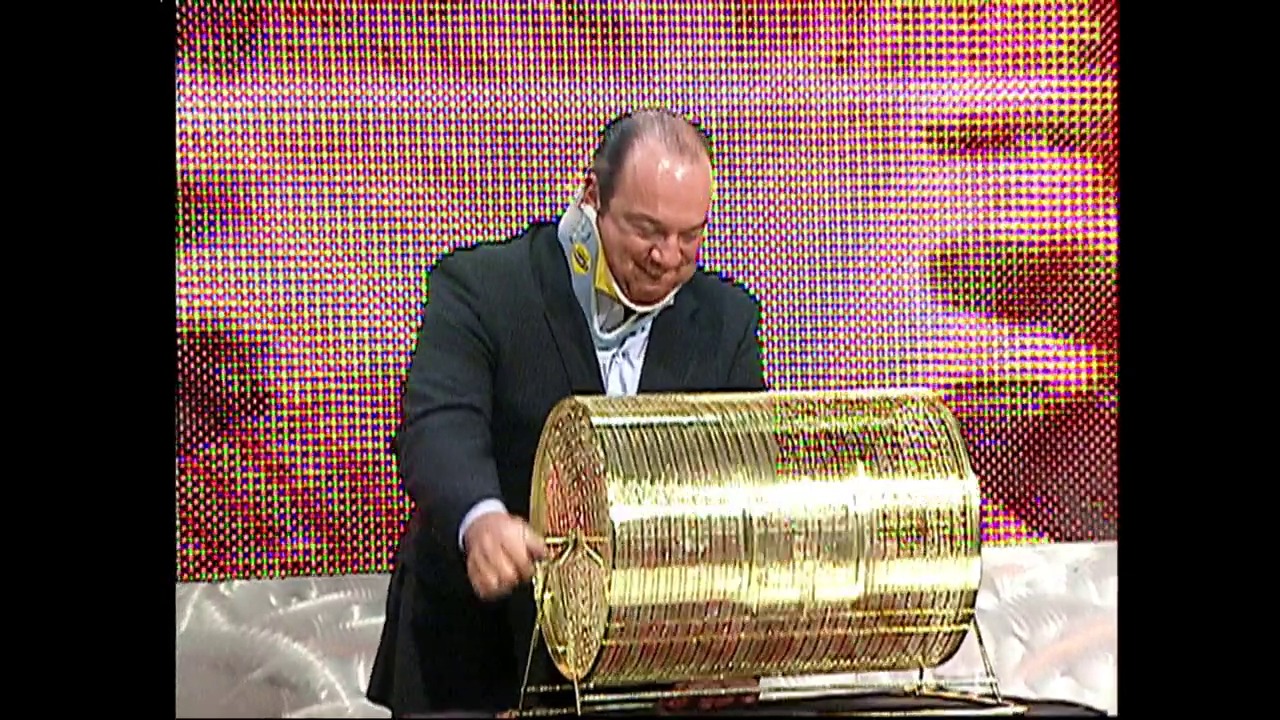 A frame from WWE SmackDown (March 25, 2004) at 0:38. A man in a dark business suit operates a large golden lottery drum against a colorful pixelated backdrop, appearing to be drawing numbers or making a selection during a WWE event broadcast.