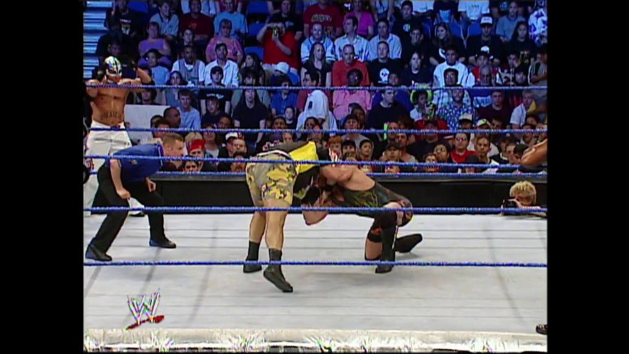 A frame from WWE SmackDown (May 6, 2004) at 1:13:58. A WWE wrestler in yellow and black attire performs a move in the ring while opponents crouch nearby. The packed crowd watches from the stands behind the blue-roped ring, with the WWE logo visible in the lower left corner.