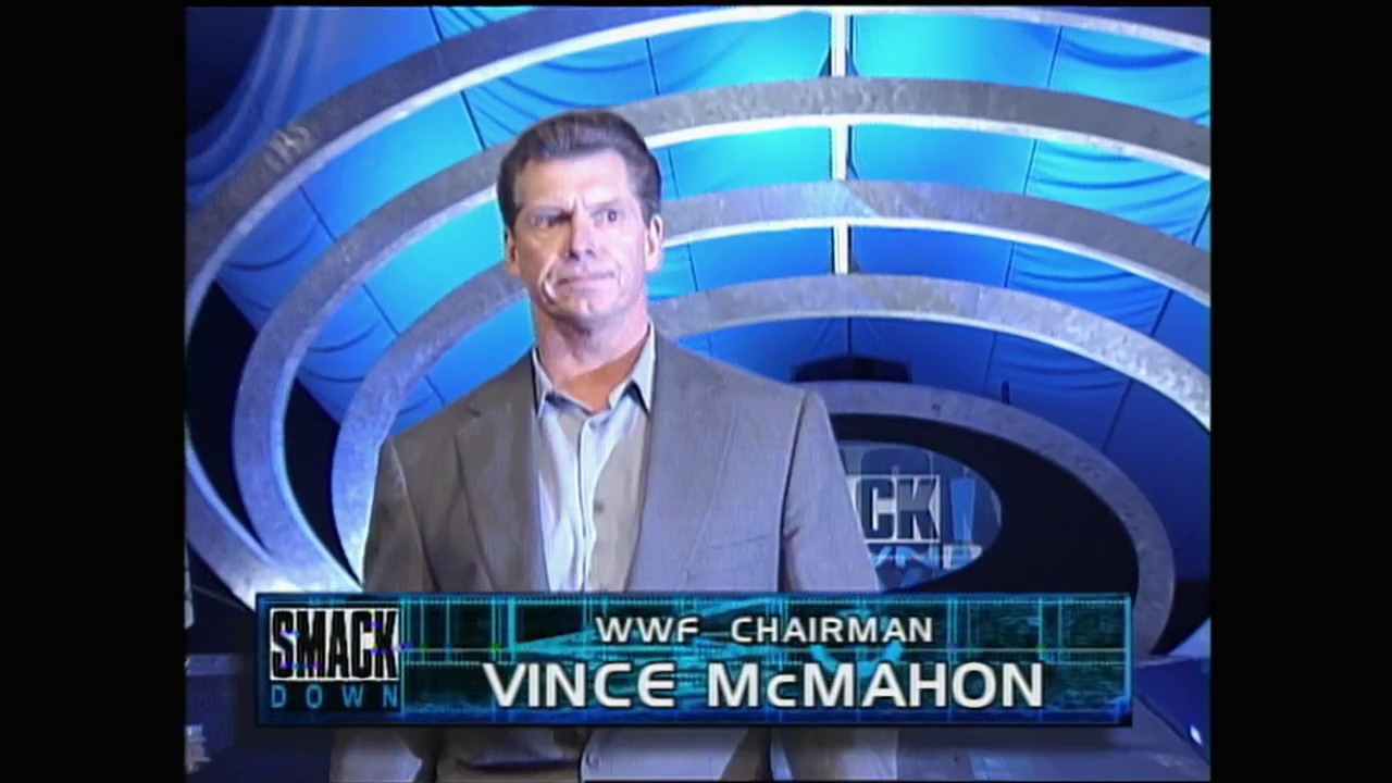 A frame from WWE SmackDown (September 23, 1999) at 1:43. A man in a tan blazer and light blue shirt stands before a blue spiral-patterned backdrop. The lower third displays "WWE SMACKDOWN" graphics and identifies him as "WWE CHAIRMAN VINCE McMAHON."
