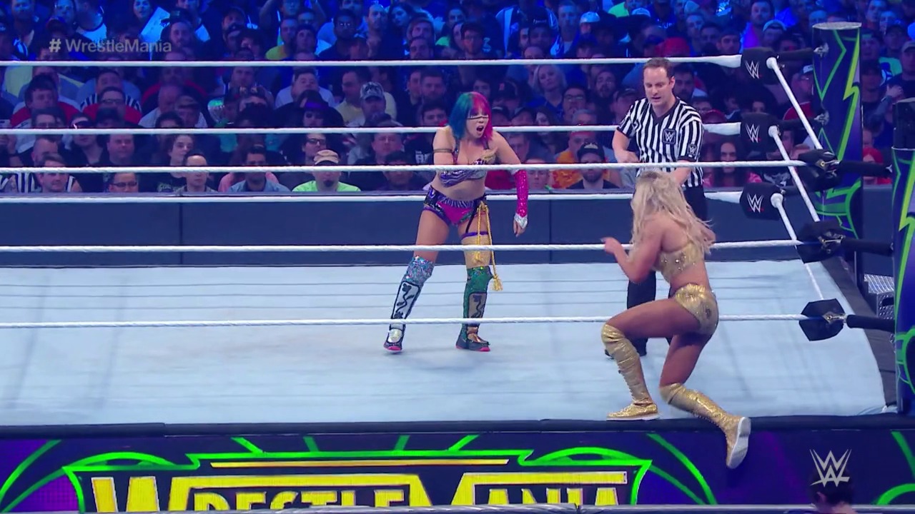 A frame from WrestleMania (2018) at 43:16. Two female wrestlers face off in a WWE ring during WrestleMania, with a referee in striped shirt overseeing the match. The crowd fills the arena in the background while bright green and yellow graphics frame the bottom of the broadcast.