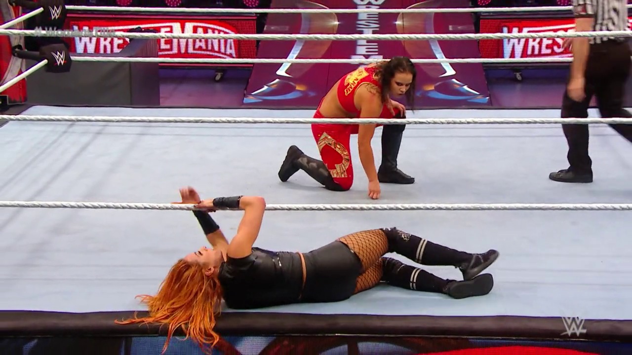 A frame from WrestleMania 36 (2020) at 1:02:25. # WrestleMania 36 Match Frame

A WWE wrestler with orange hair lies on the mat in black attire, while an opponent in red stands nearby on one knee. The WrestleMania branding is visible on the ring backdrop with purple and red lighting.