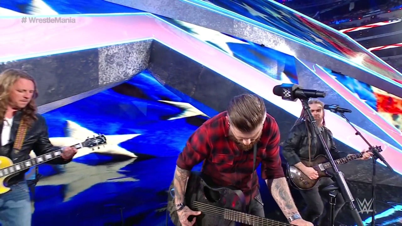 A frame from WrestleMania 38 (2022) at 2:47. A rock band performs on the WrestleMania stage, with three guitarists in focus playing against a dynamic blue and pink backdrop featuring the WWE logo and lighting effects.