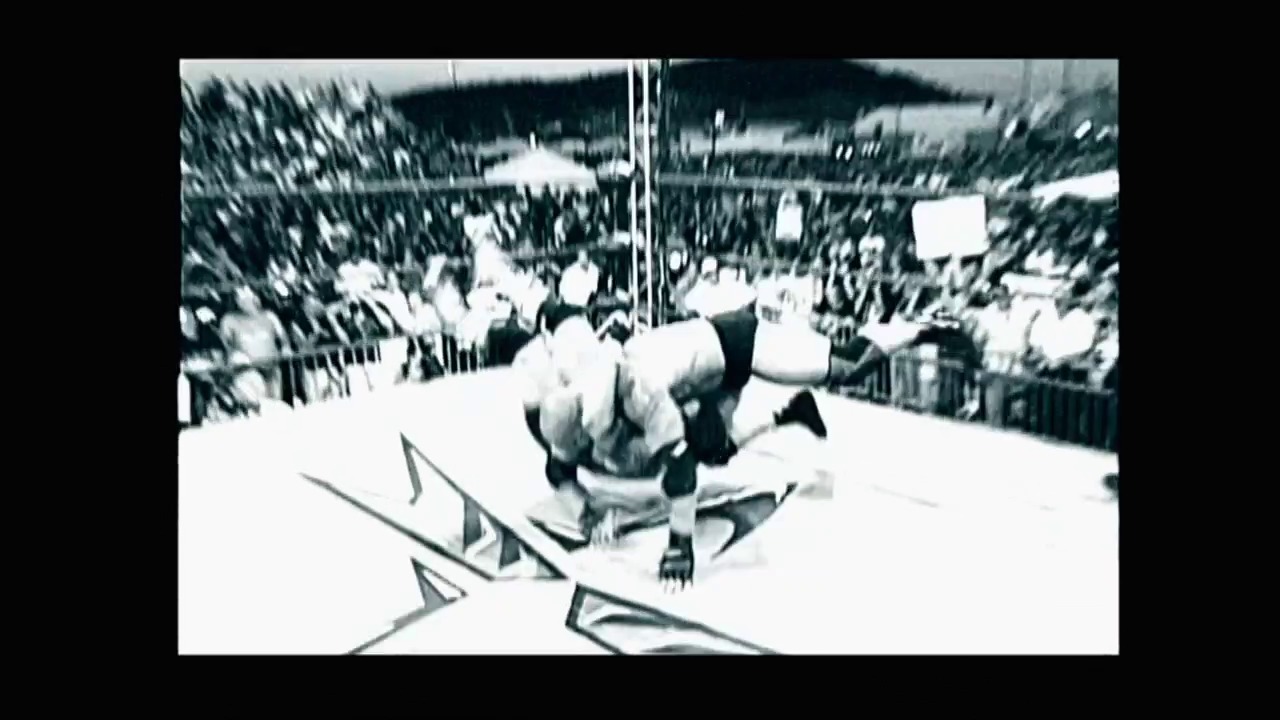 A frame from WWE Backlash (2003) at 0:27. A wrestling match is underway in a packed arena, with wrestlers grappling in the ring while a large crowd fills the stands. The black and white footage shows the intensity of the moment, with graphics visible at the bottom of the frame.