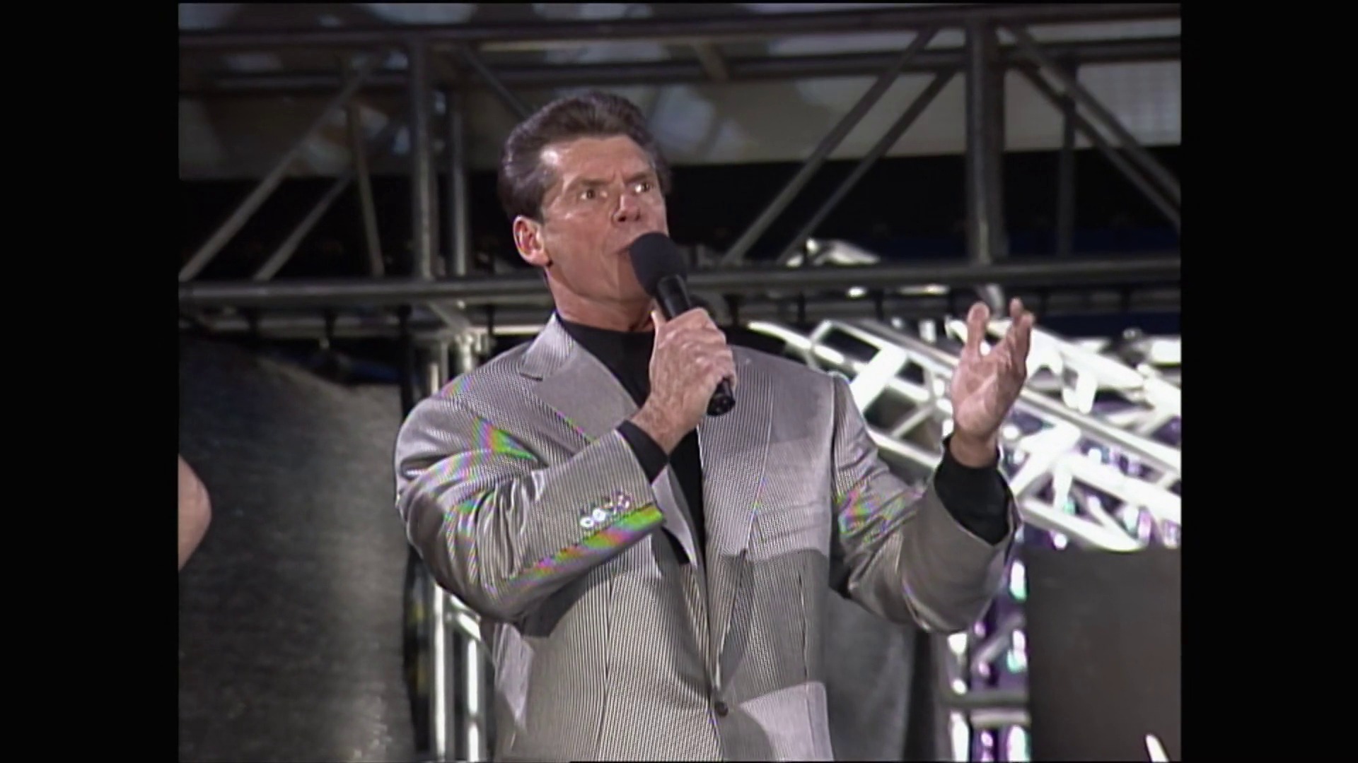 A frame from WWE Raw (February 13, 1999) at 13:49. A man in a gray pinstriped jacket with colorful shoulder designs holds a microphone while speaking in a wrestling arena, with the ring and steel structure visible in the background.