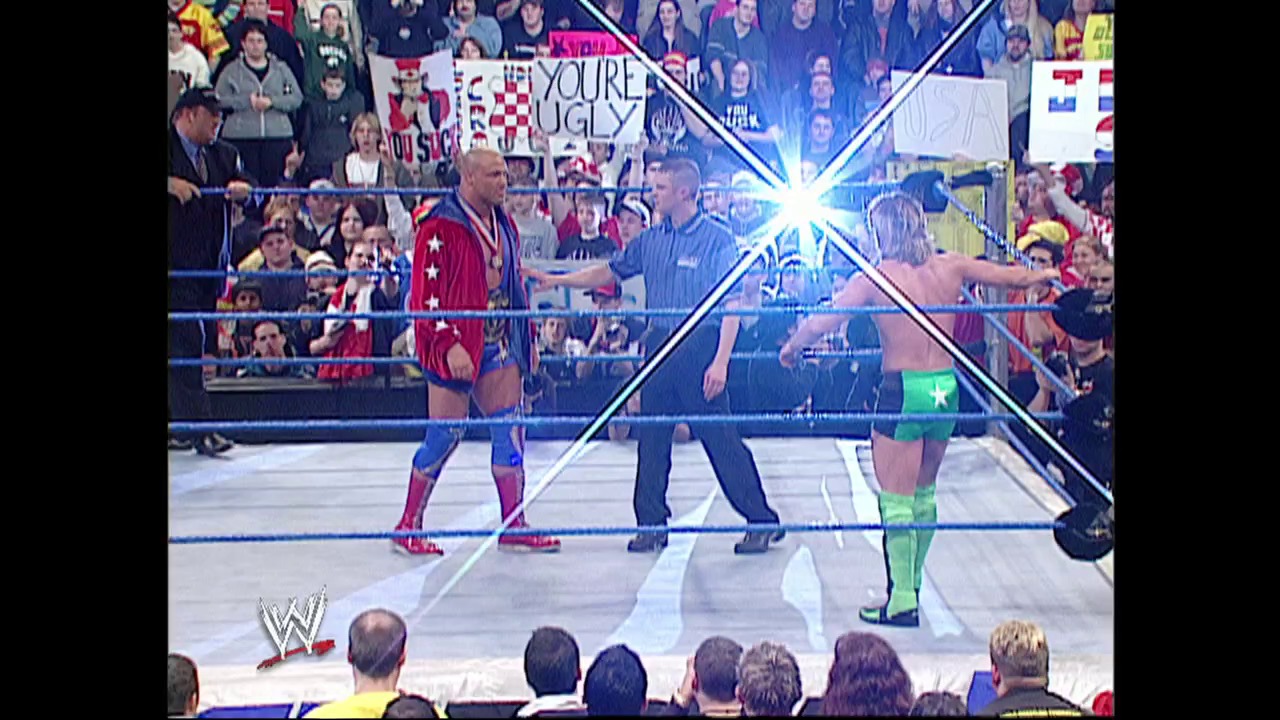 A frame from WWE SmackDown (February 27, 2003) at 50:18. A WWE referee stands between two wrestlers in the ring—one in red and blue attire on the left, another in green and black on the right. The crowd fills the arena behind them as blue pyrotechnic effects light up the stage.